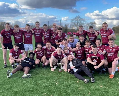 A group of males wearing burgundy shirts stand and sit around a silver trophy on grass.